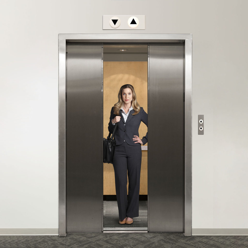 A businesswoman holding a shoulder bag, looks directly at the camera as the doors on the elevator are about to close on her.  The arrow indicates that the elevator is moving to a higher floor.